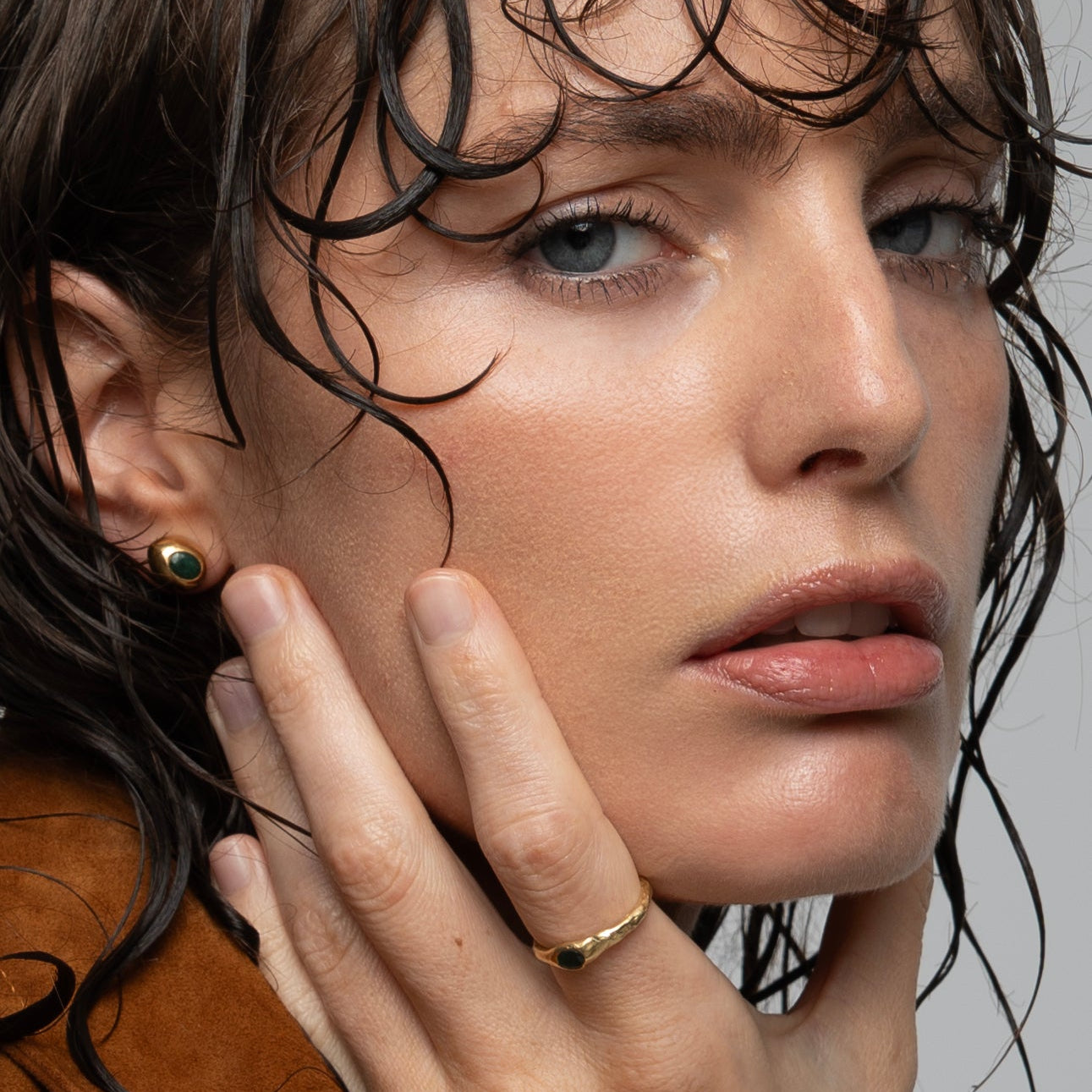 Woman wearing gold earrings and a ring, with wet hair against a neutral background